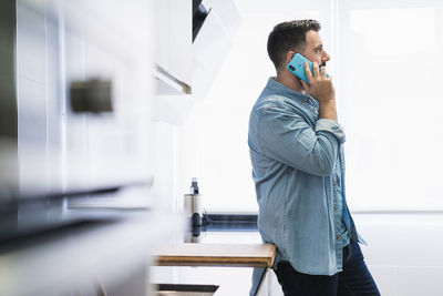 Man cooking crepes in the kitchen with a mobile phone in a denim shirt