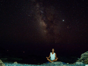 Woman meditating while sitting on rock against sky at night