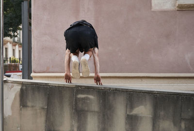 Back view of concentrated male jumping over stone fence and balancing on arm while performing stunt and doing parkour