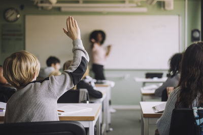 Rear view of student raising hand while teacher teaching in classroom