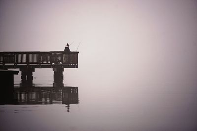 Rear view of person on pier fishing by lake during foggy weather