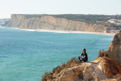 Man sitting on cliff by sea against clear sky