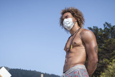 Shirtless young man wearing face mask while standing against clear blue sky during covid-19