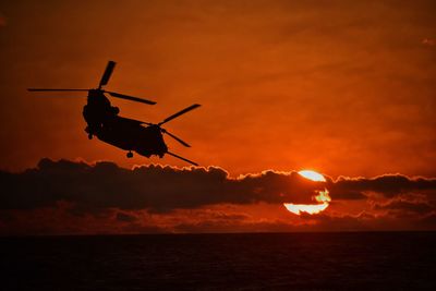 Silhouette helicopter flying over sea against sky during sunset