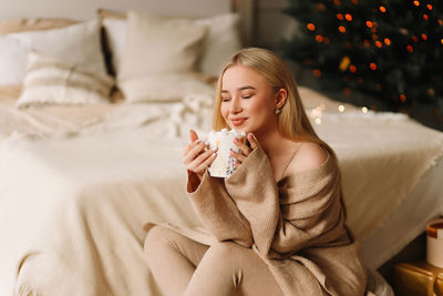 A beautiful young woman in a cardigan holds a mug drinking coffee while enjoying a holiday at home