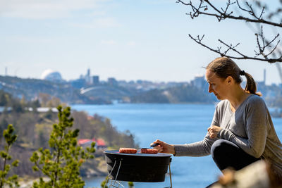 Side view of woman preparing meat on barbecue by river