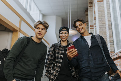 Low angle view of smiling male friends with mobile phone while walking in school corridor