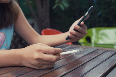 Midsection of woman with cigarette using mobile phone at table