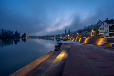 Illuminated buildings by river against sky at dusk