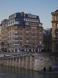 People on canal by buildings in city against clear sky