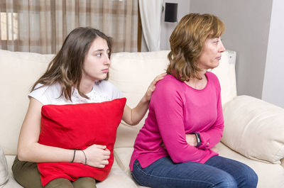 Daughter holding cushion comforting angry mother on sofa at home