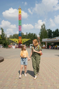 Mother and daughter eating tasty cold sweet ice creams while walking holding hands in public amusement park on summer day in city