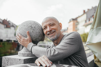 Cheerful senior man with a beard hugging a large stone ball in an urban park setting