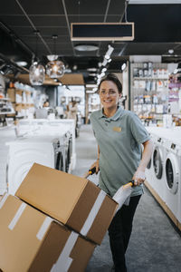 Portrait of smiling saleswoman carrying boxes in push cart at electronics store