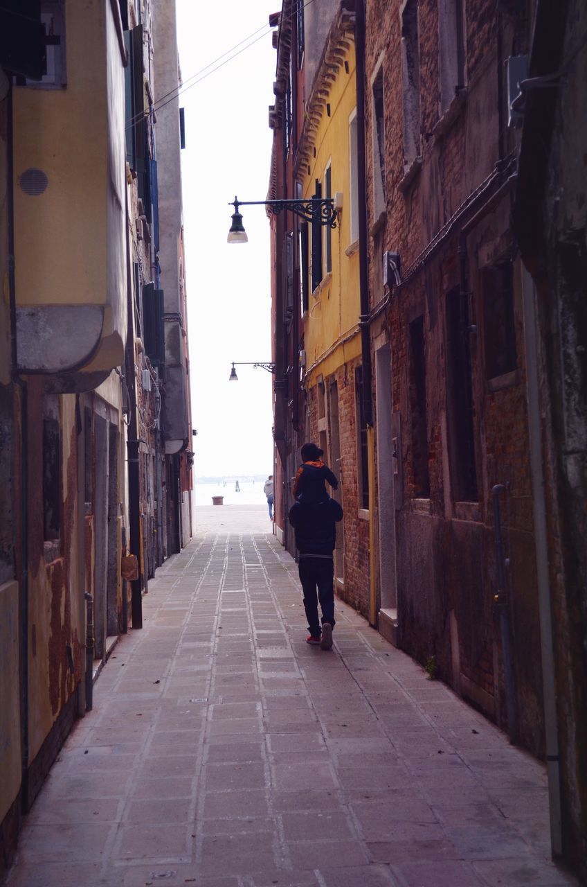 REAR VIEW OF MAN WALKING ON FOOTPATH AMIDST BUILDINGS IN CITY