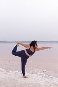 Side view of curvy female in sportswear balancing in natarajasana on shore of pink pond and practicing yoga in summer