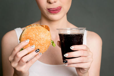Midsection of woman holding burger and cold drink against gray background