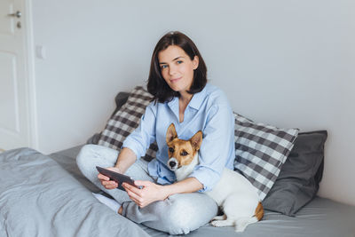 Woman with dog using digital tablet while sitting on bed against wall at home