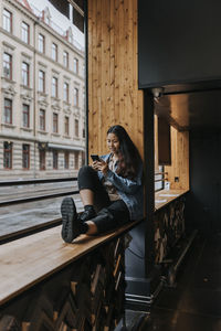 Full length of female store owner using smart phone sitting on window sill