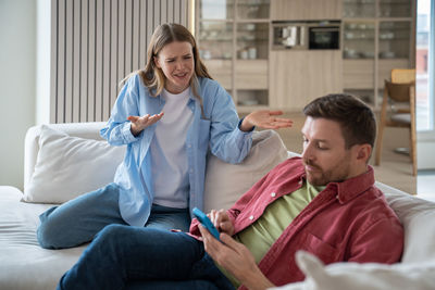 Nervous reproachful wife shouting at calm neglecting husband sitting on sofa, playing mobile games