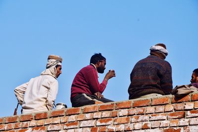 Low angle view of people sitting on retaining wall against clear sky