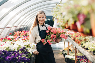 A cheerful woman in an apron holds a pot of flowers in her hands while working in a greenhouse. 