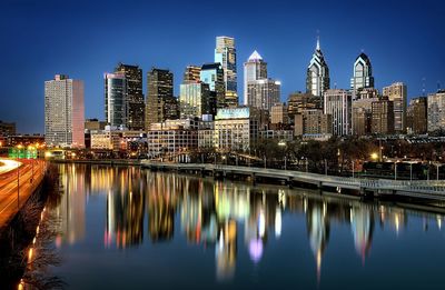 Modern illuminated buildings by river against clear sky in city at night