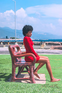 Side view of woman sitting on bench at beach against sky and mountain