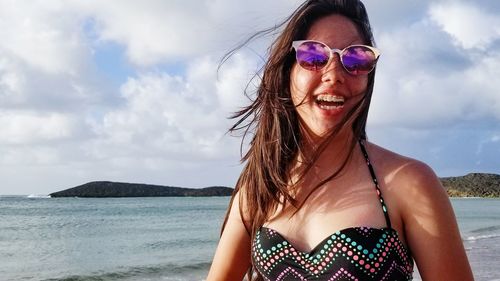 Portrait of cheerful teenage girl wearing sunglasses at beach against sky