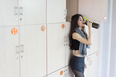 Woman drinking water from bottle while standing against locker