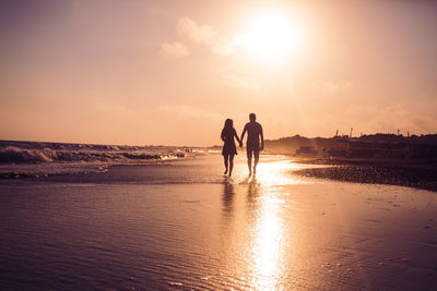 Silhouette couple walking on beach against sky during sunset
