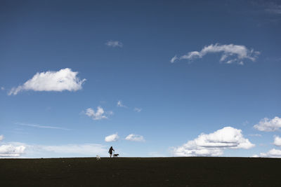 Mid distance view of male hiker with dogs walking on landscape against blue sky