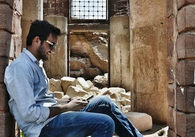 Side view of young man using smart phone while sitting at ruins