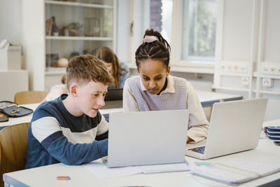 Boy sharing laptop with female friend sitting at desk in classroom