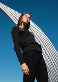 Low angle view of woman by metallic structure against clear blue sky