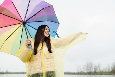 Beautiful smiling brunette woman in yellow raincoat holding rainbow umbrella out in the rain