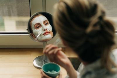 A woman applies a clay mask to her face while looking in the mirror.
