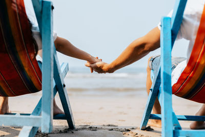Cropped image of couple holding hands while relaxing on chair at beach