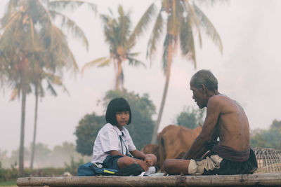 Portrait of girl sitting with shirtless grandfather against trees