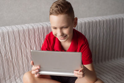 A cute boy in a red t-shirt is sitting playing on a tablet and laughing
