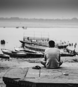 Rear view of man sitting on pier with boats in background