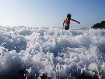 Rear view of shirtless boy standing in sea against sky during sunny day