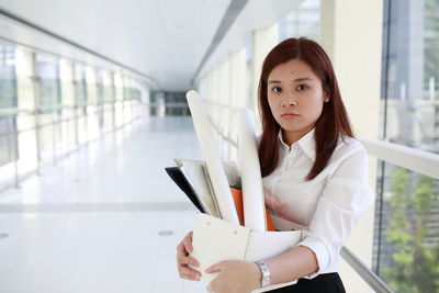 Portrait of young woman holding box while standing in office corridor