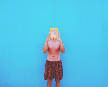 Shirtless man holding picture frame in front of face against blue wall