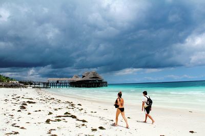 Full length of woman in bikini walking with man at beach against cloudy sky