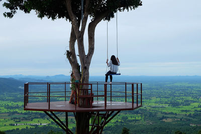 Rear view of woman sitting on swing by tree against sky