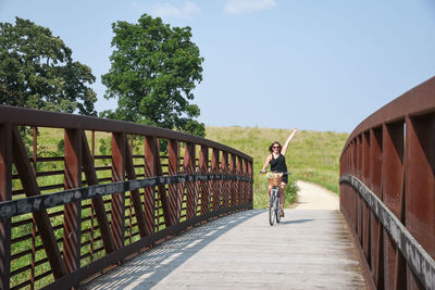 Full length of mature woman riding bicycle on footbridge against blue sky during sunny day