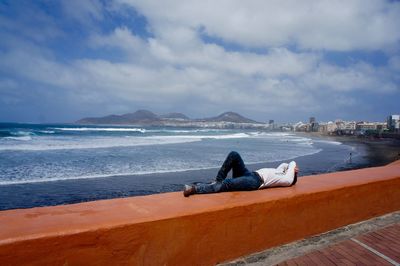 Man lying on retaining wall by sea against cloudy sky