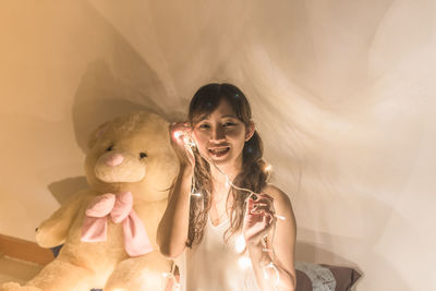 Portrait of young woman with illuminated string light sitting by wall at home