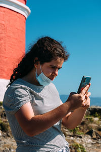 Young woman wearing face mask using mobile phone against sky